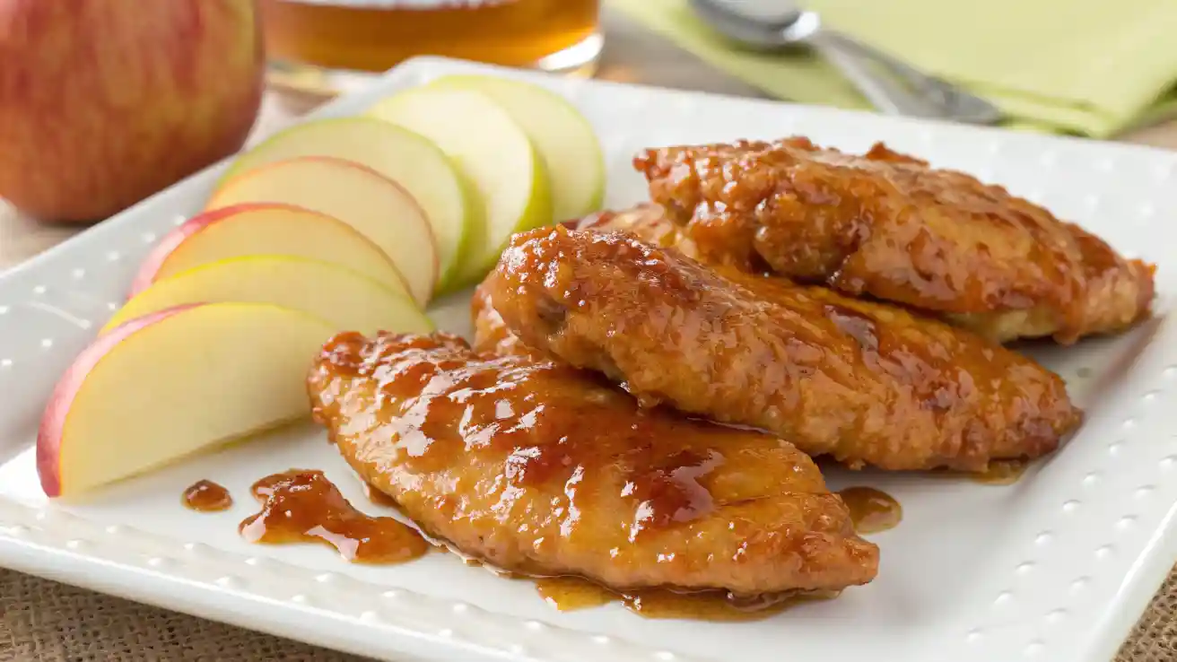 Close-up of golden-brown Apple and Honey-Glazed Chicken Tenders on a white plate with a shiny glaze and apple slices.