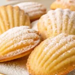 Golden-brown Madeleine cookies on a white plate, some dusted with powdered sugar and others dipped in chocolate, styled with lemon zest and a cup of tea.