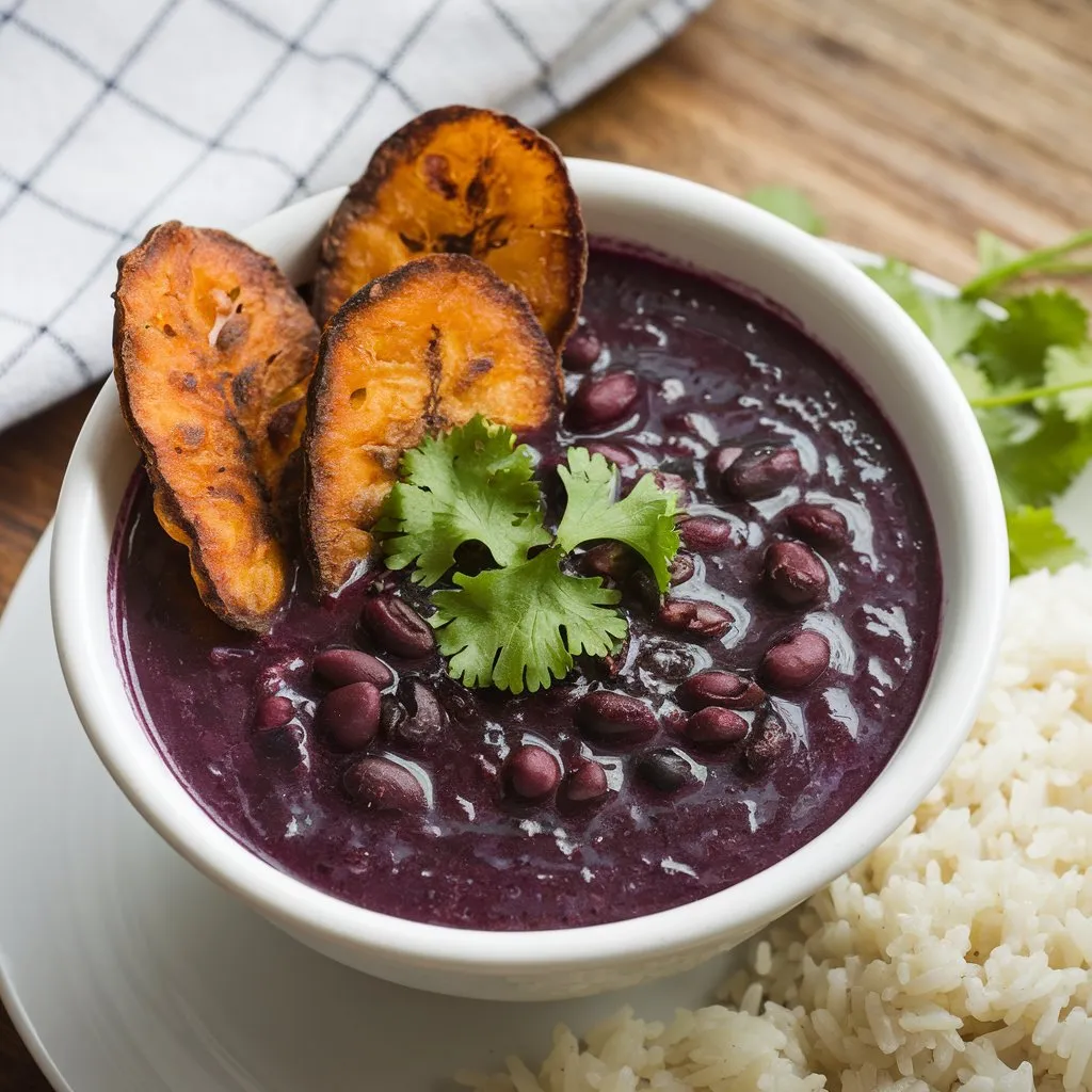 A photo of a rich purple black bean soup served with crispy fried plantains and a side of white rice. Fresh cilantro is sprinkled on top of the soup, placed on a simple wooden table with a checkered cloth.