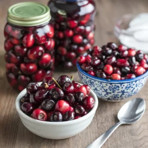 A photo of a recipe featuring canned cherries and cranberries. In the foreground, a bowl of mixed cherries and cranberries alongside a bowl of canned cherries and cranberries, with a spoon resting beside them. In the background, two jars of cherries and one jar of cranberries are arranged on a wooden surface.