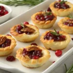 Top-down view of cranberry Brie bites with puff pastry, cranberry sauce, rosemary, and pecans on a white platter with scattered cranberries.