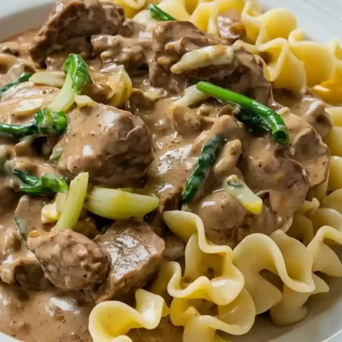 A serving of beef stroganoff with noodles, creamy sauce, chunks of beef, mushrooms, and onions on a white plate.