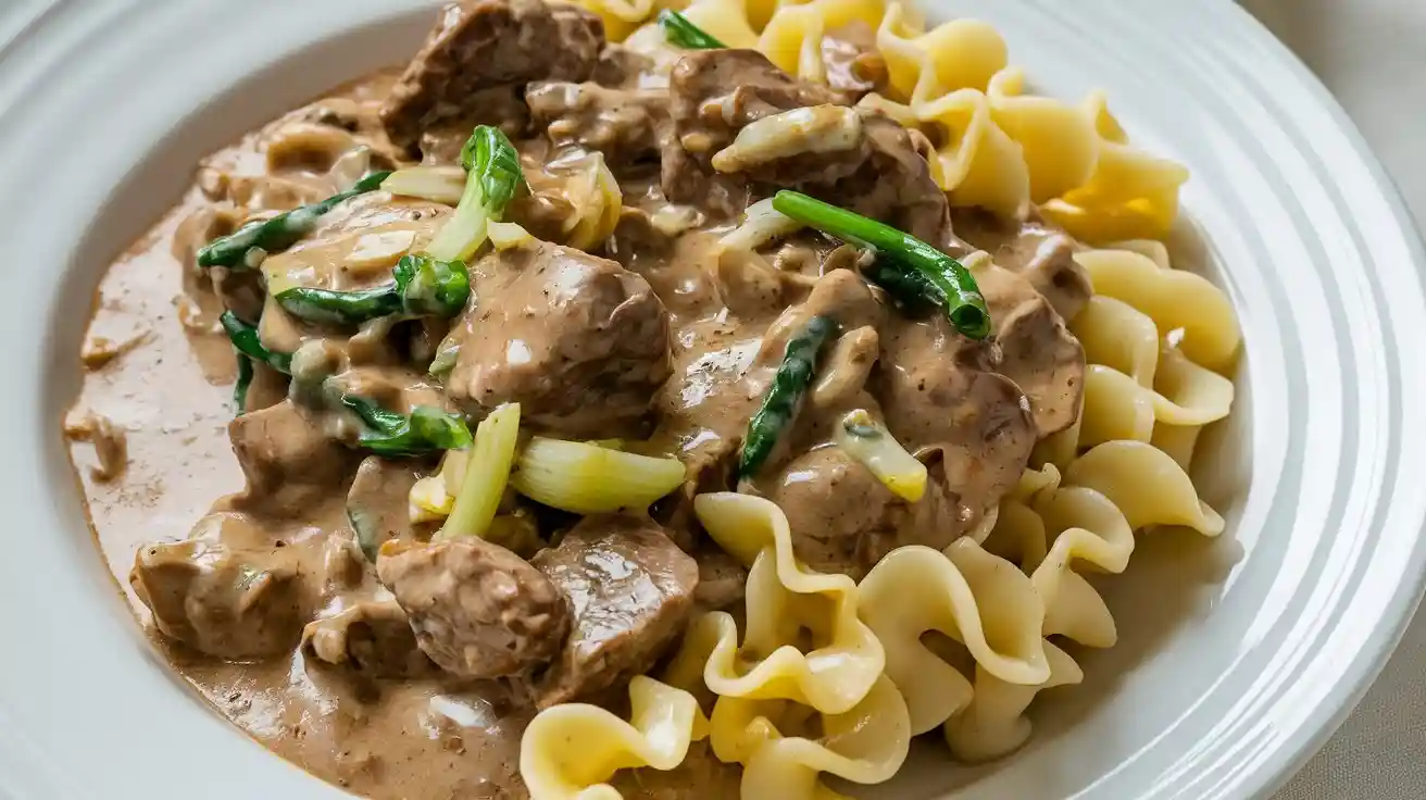 A serving of beef stroganoff with noodles, creamy sauce, chunks of beef, mushrooms, and onions on a white plate.