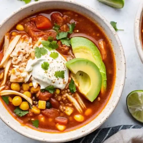 Overhead close-up of chicken taco soup in a rustic bowl, topped with avocado, cheese, jalapeños, sour cream, cilantro, and lime.