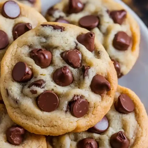 Close-up of a plate of golden brown chocolate chip cookies with crispy edges and plenty of chocolate chips, set on a wooden surface.