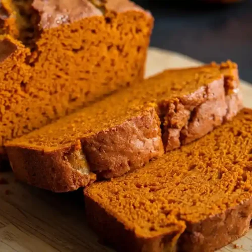 Moist pumpkin bread slices on a wooden board, showcasing a delicious pumpkin bread recipe, with the loaf in a pan in the background.