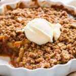 Overhead shot of a golden apple crumble in a round white dish, topped with vanilla ice cream and a cinnamon oat topping.