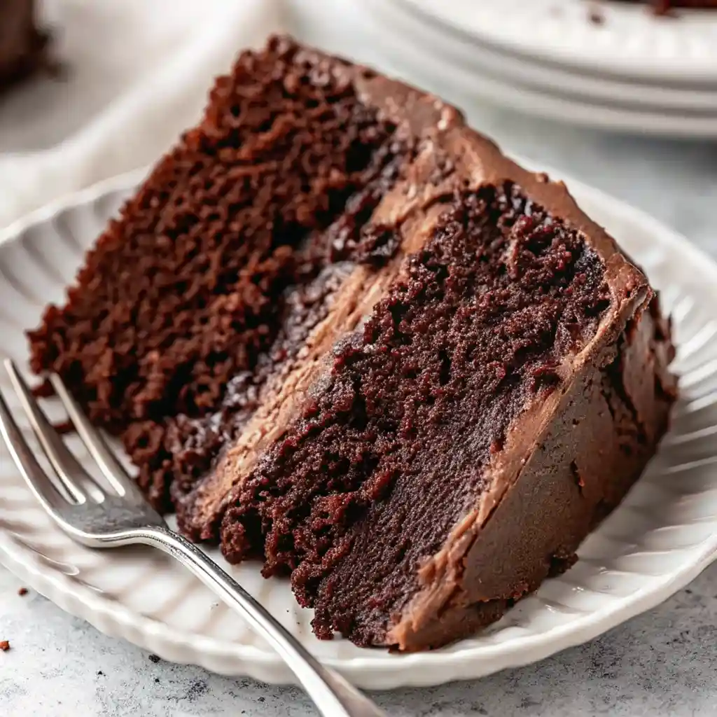 a slice of moist chocolate layer cake with buttercream frosting on a white ceramic plate and fork.
