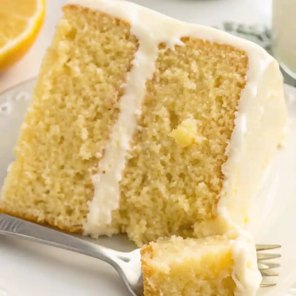 Close-up of a single slice of two-layer lemon cake with creamy frosting and a fork holding a bite, set on a white dessert plate.