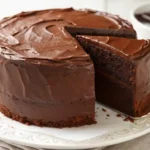 a moist Chocolate Cake Recipe with rich frosting and one slice removed on a white plate lined with parchment paper.