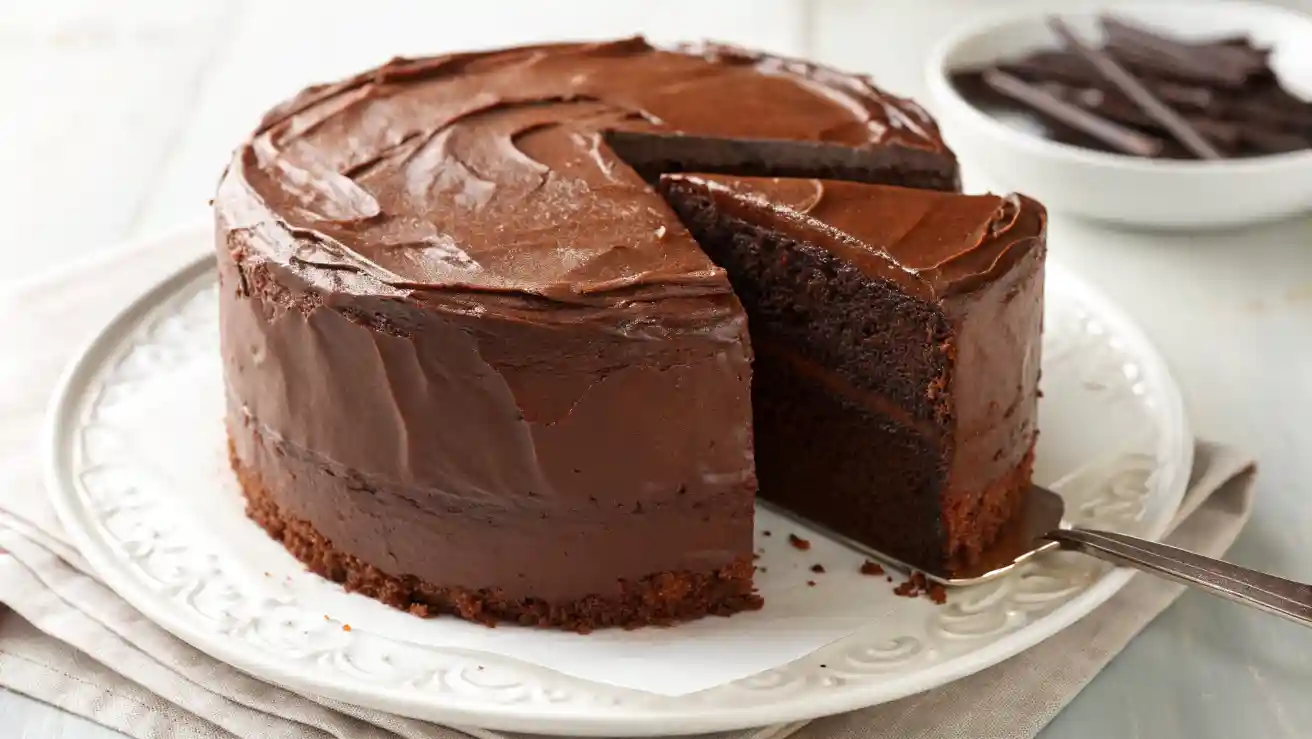 a moist Chocolate Cake Recipe with rich frosting and one slice removed on a white plate lined with parchment paper.