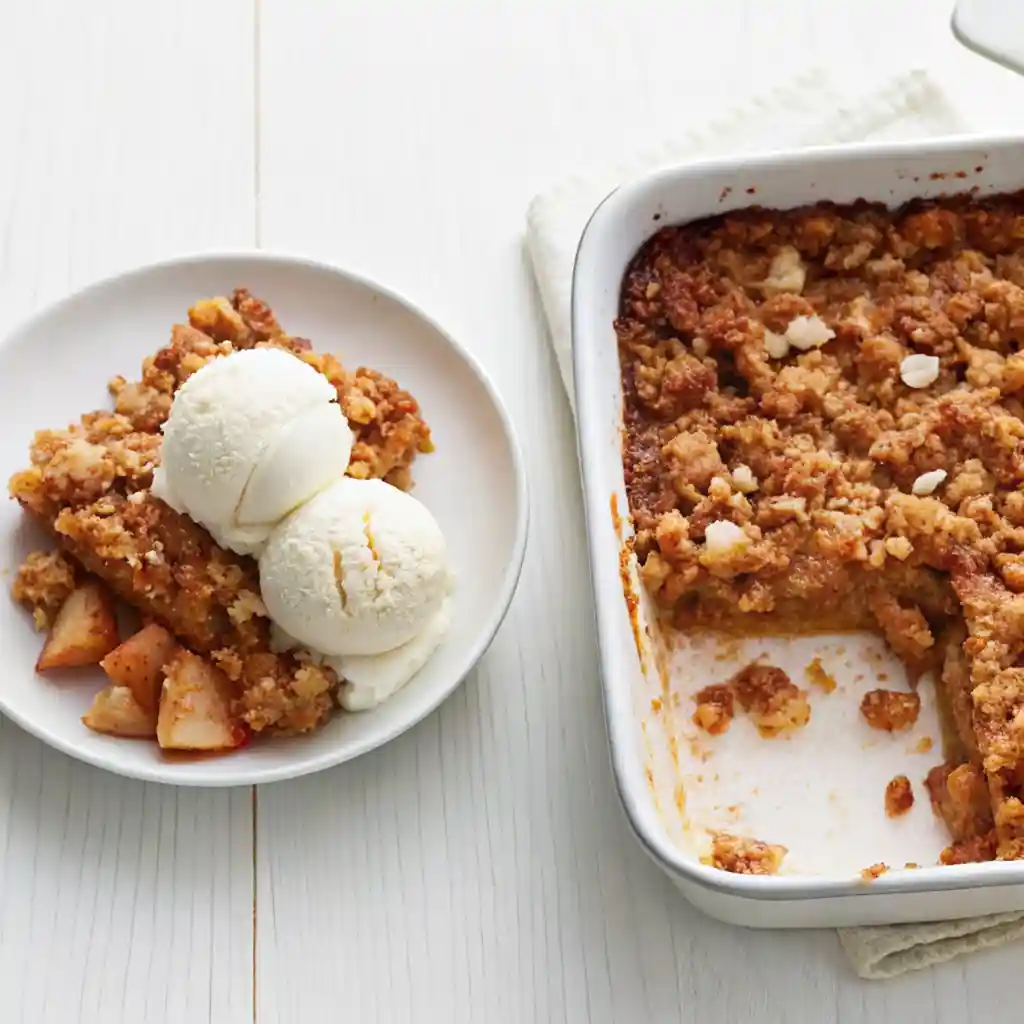 Overhead view of apple crumble in a white dish with a serving removed and plated with melting vanilla ice cream.