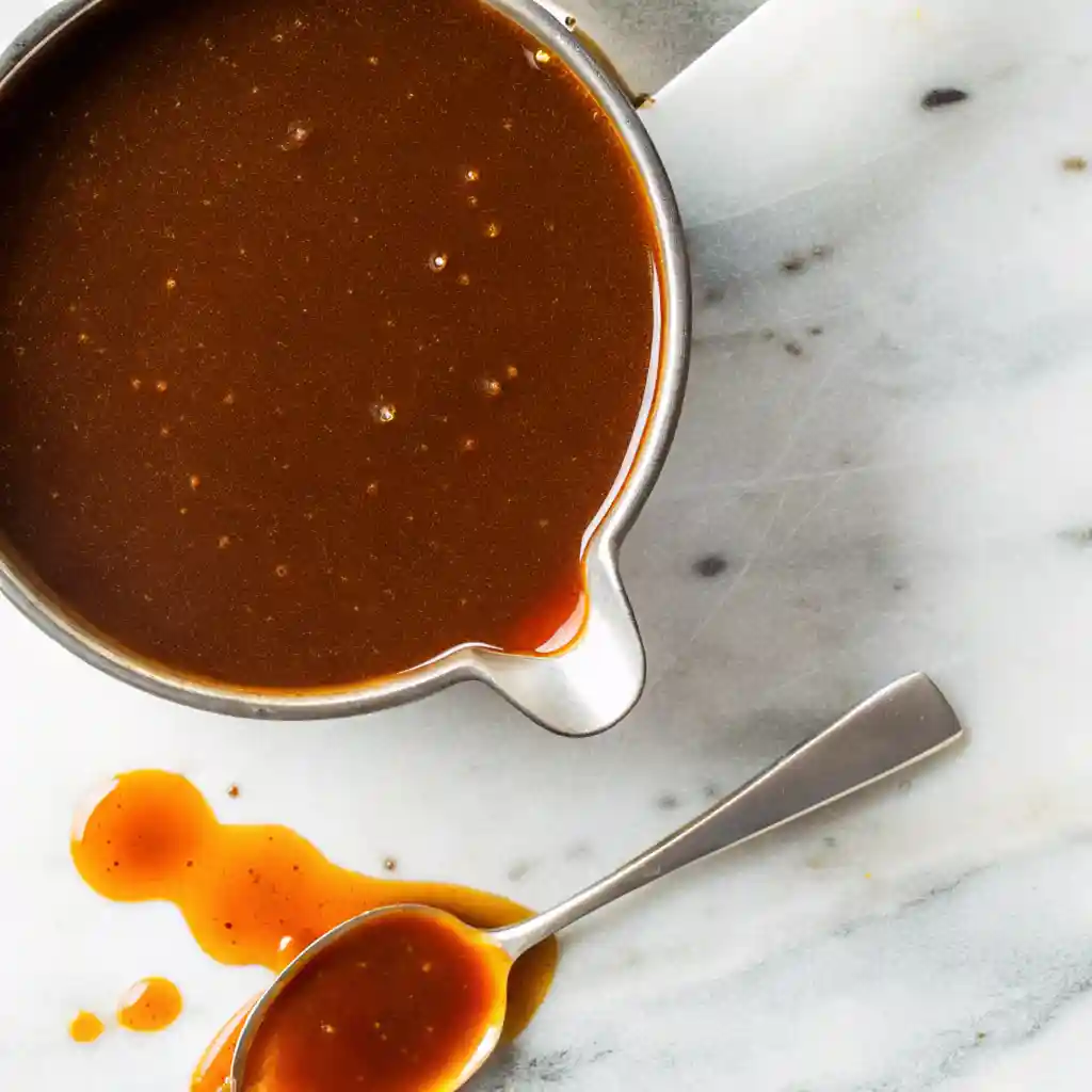 Overhead view of a metal saucepan filled with glossy demi-glace, with a spoon and sauce drips on white marble.
