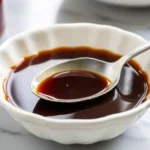 Close-up of a white bowl filled with glossy demi-glace, with a silver spoon resting inside the rich brown sauce.