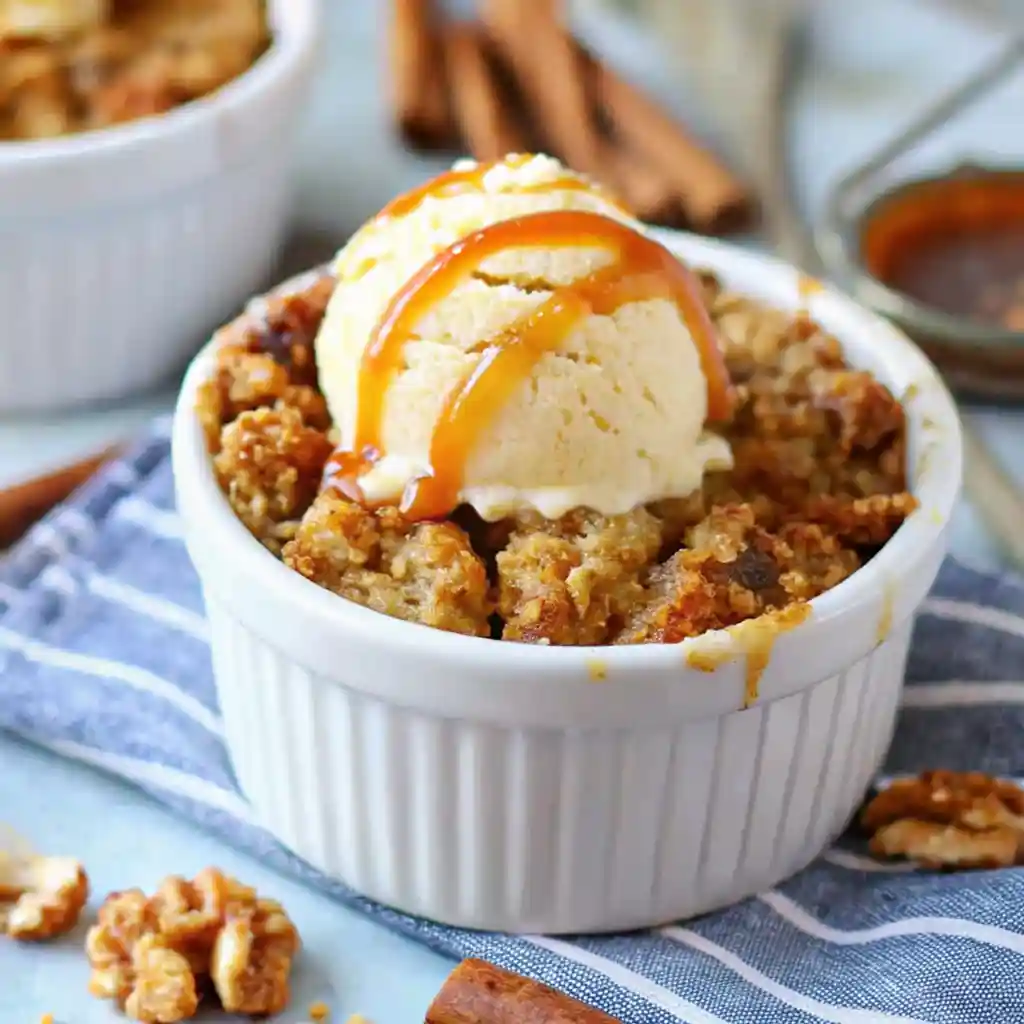 Single-serving apple crumble in a white ramekin with ice cream and caramel drizzle on a striped cloth with cinnamon and walnuts.
