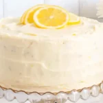 Two-layer lemon cake on a clear glass stand, frosted with lemon zest buttercream and topped with lemon slices, surrounded by lemons and a blue checkered napkin.