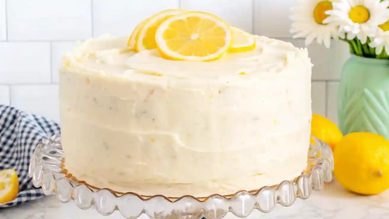 Two-layer lemon cake on a clear glass stand, frosted with lemon zest buttercream and topped with lemon slices, surrounded by lemons and a blue checkered napkin.