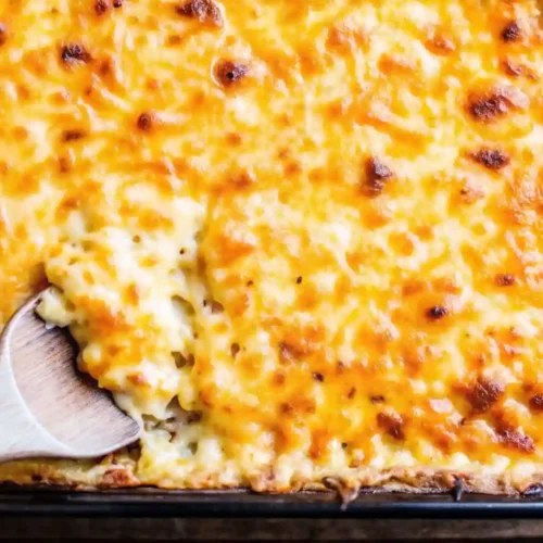 Creamy baked mac and cheese in a casserole dish with a golden cheese crust and a wooden spoon scooping out a portion
