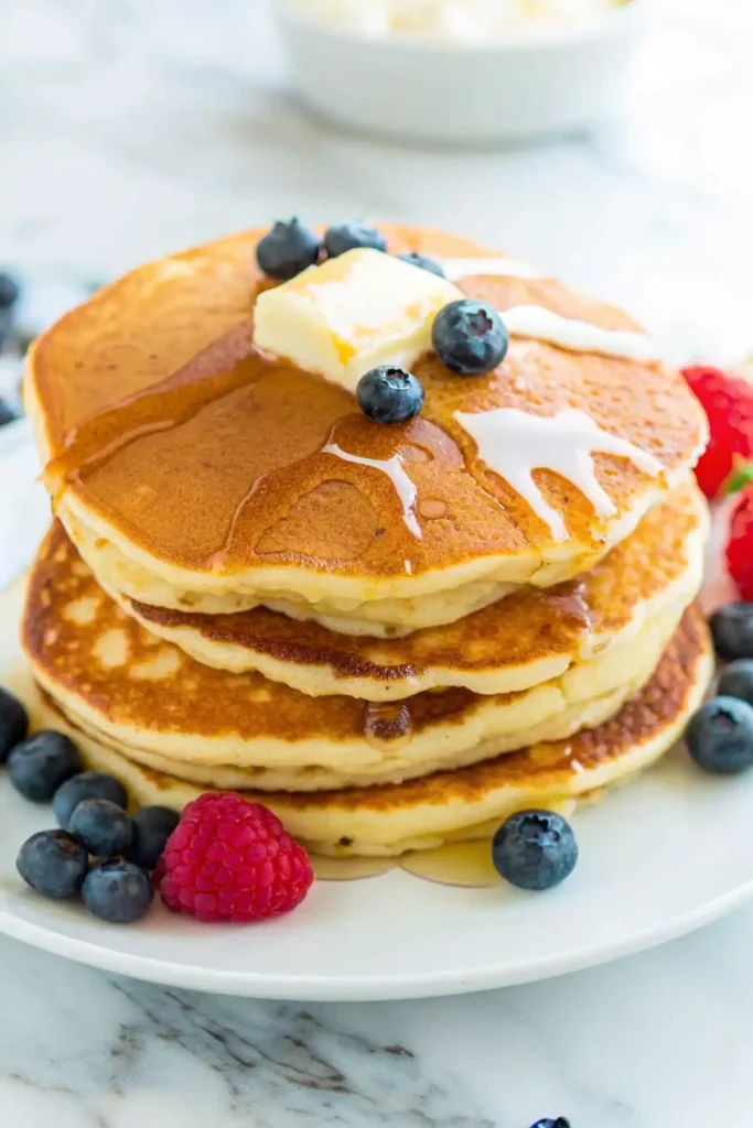 Stack of fluffy pancakes topped with butter syrup and fresh blueberries on a white plate.