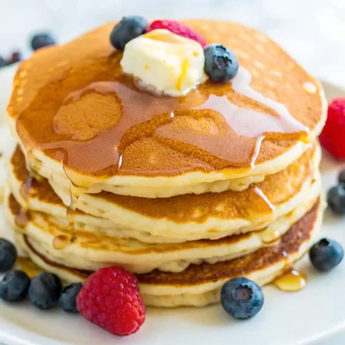 Stack of fluffy golden pancakes topped with butter, syrup and fresh berries on a white plate.
