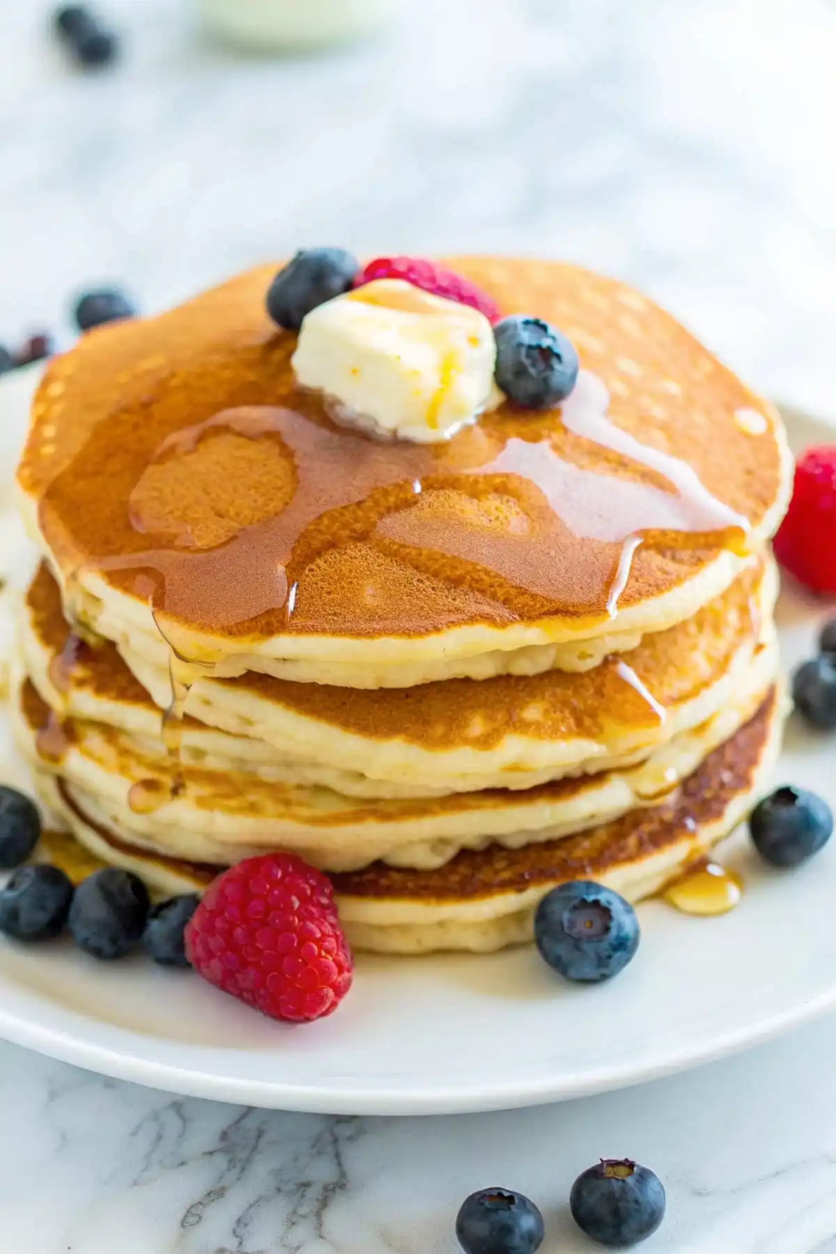 Stack of fluffy golden pancakes topped with butter, syrup and fresh berries on a white plate.