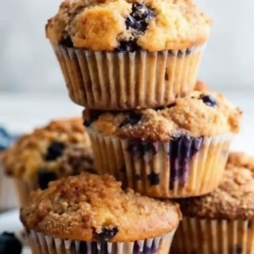 Freshly baked blueberry muffins with crumb topping stacked on a white surface, surrounded by loose blueberries.