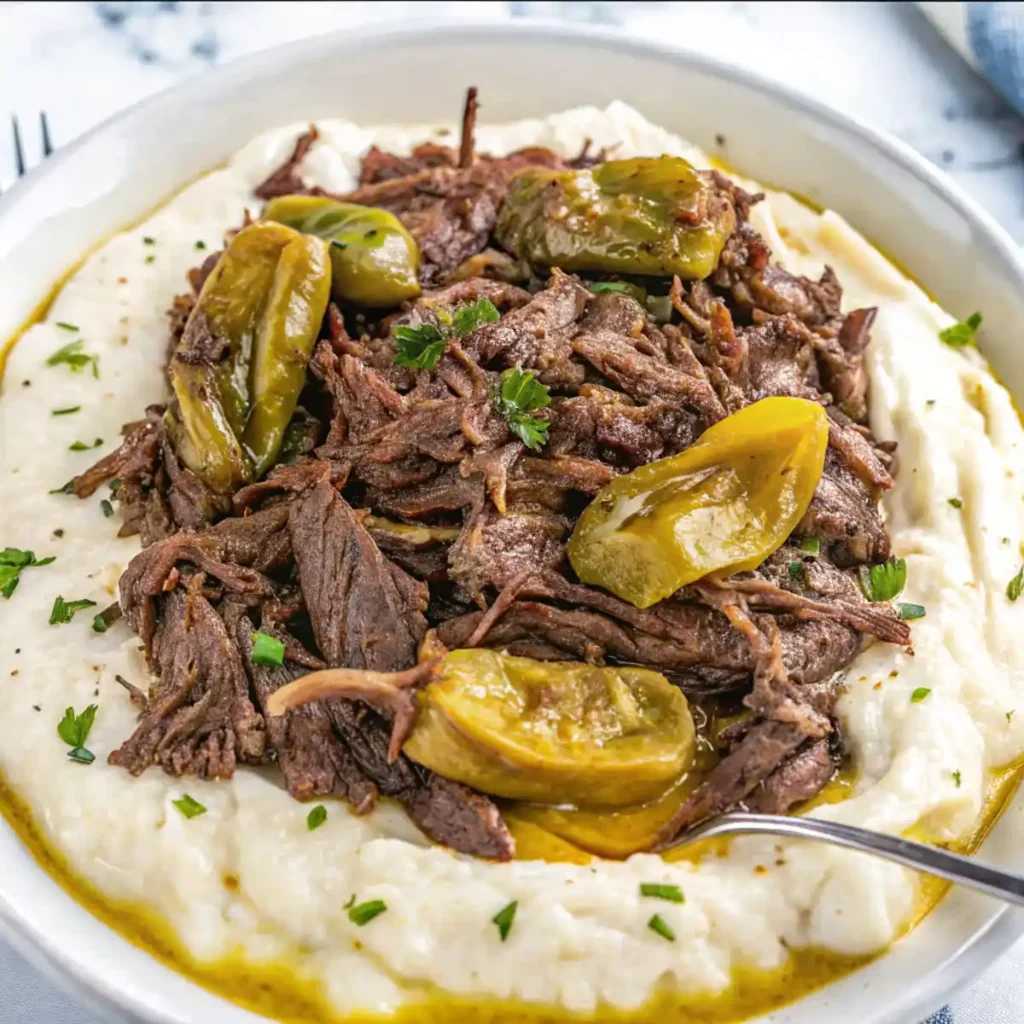 Crockpot Mississippi pot roast with pepperoncini peppers served over creamy mashed potatoes in a white bowl, garnished with parsley.