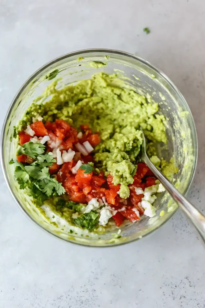 Glass bowl of partially mixed guacamole with avocado, diced tomato, onion, cilantro, and crumbled cheese