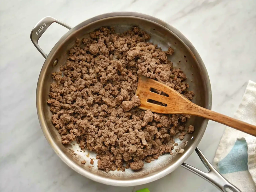 Cooked ground beef in a stainless steel skillet with a wooden spatula on a light marble surface.