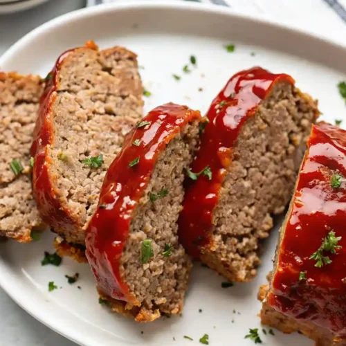 Sliced homemade meatloaf with glossy red glaze, served on a white platter and garnished with chopped parsley.