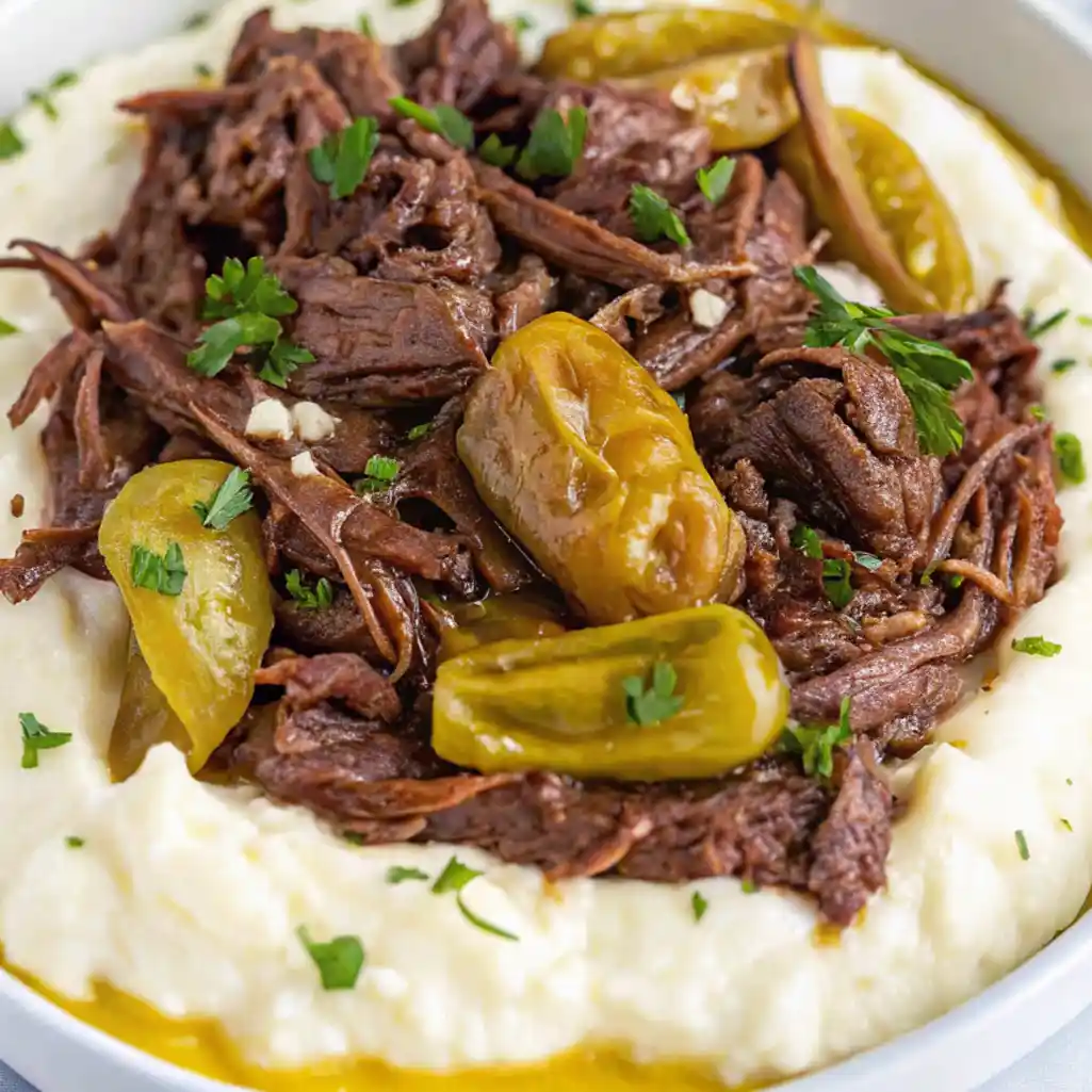 Shredded Mississippi pot roast with pepperoncini peppers served over mashed potatoes and garnished with fresh parsley in a white bowl.