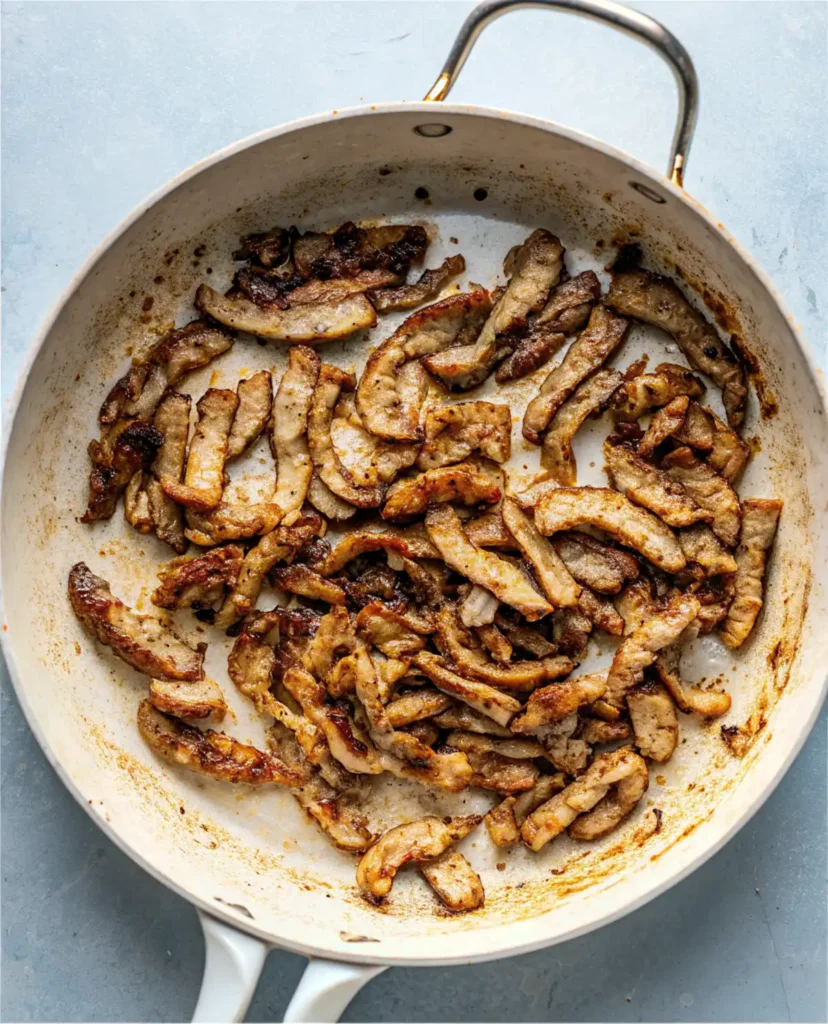 Cooked beef strips in a white skillet with browned edges and seared marks.