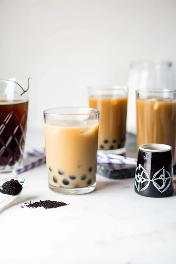 A glass of iced milk tea with tapioca pearls on a white surface with brewed tea and toppings in the background.