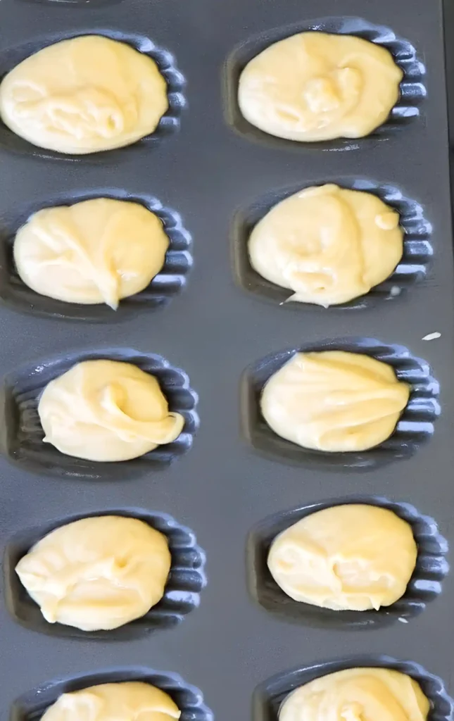 Madeleine batter scooped into a dark nonstick pan with shell shaped molds.