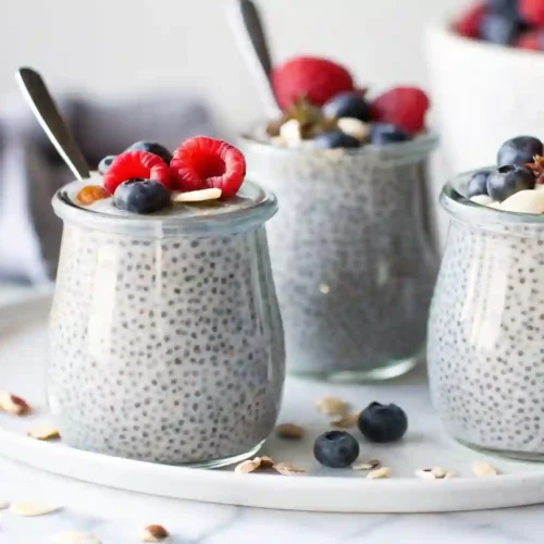 Three glass jars of chia seed pudding topped with fresh raspberries, blueberries, and nuts on a white tray.