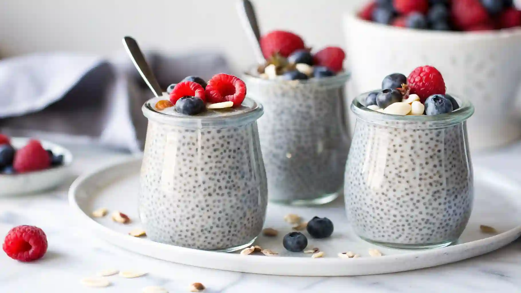 Three glass jars of chia seed pudding topped with fresh raspberries, blueberries, and nuts on a white tray.