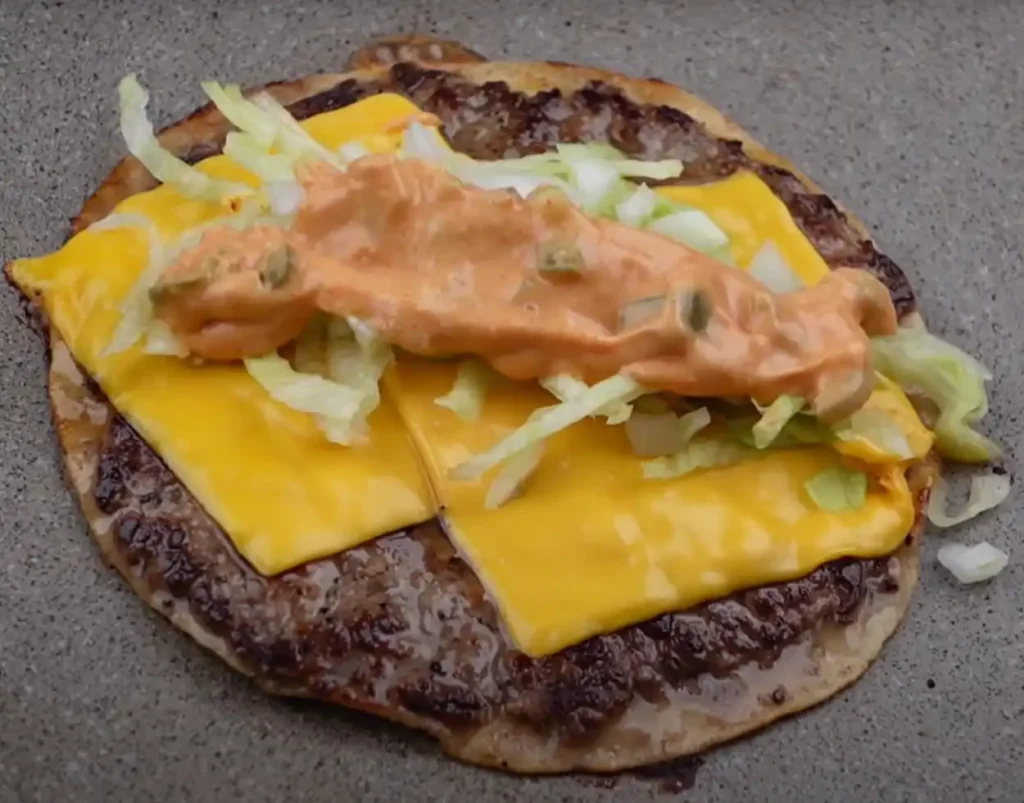 Cheddar cheese melting on a smash burger patty with lettuce, onions, and burger sauce on top of a toasted tortilla.