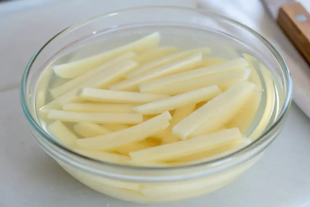 Raw potato sticks soaking in water in a clear glass bowl during French fry preparation.