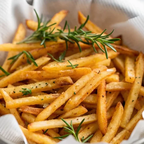 Crispy homemade French fries topped with fresh rosemary in a white paper-lined bowl.