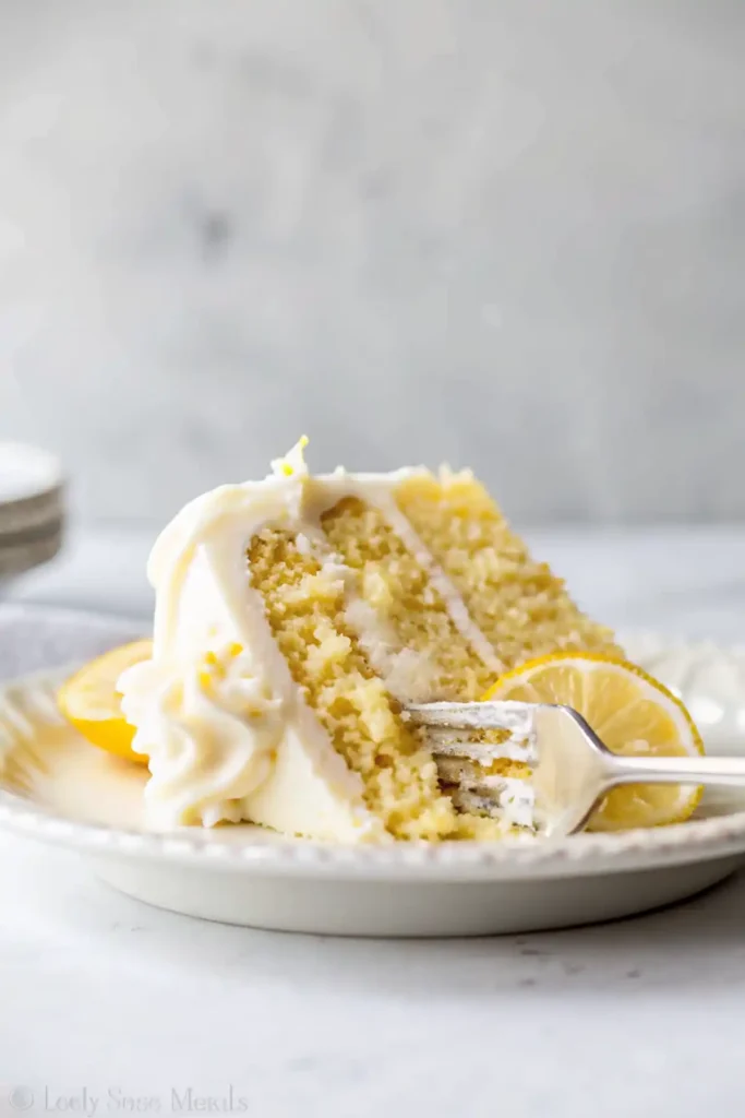 Slice of moist lemon cake with creamy frosting on a plate and a fork taking a bite.