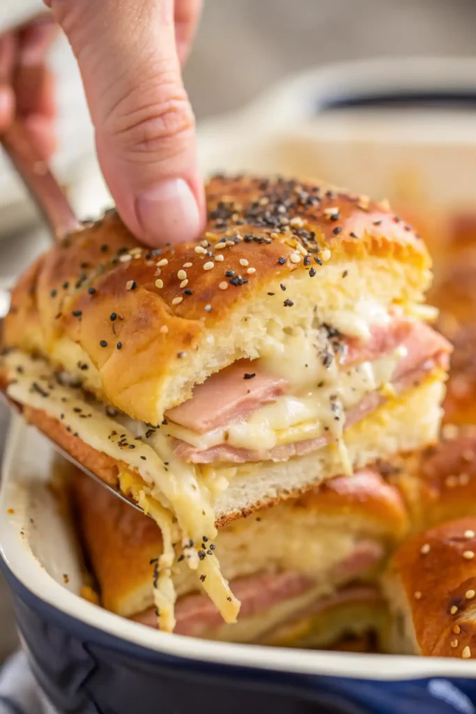 Cheesy ham and cheese slider being lifted from a baking dish with melted cheese stretching