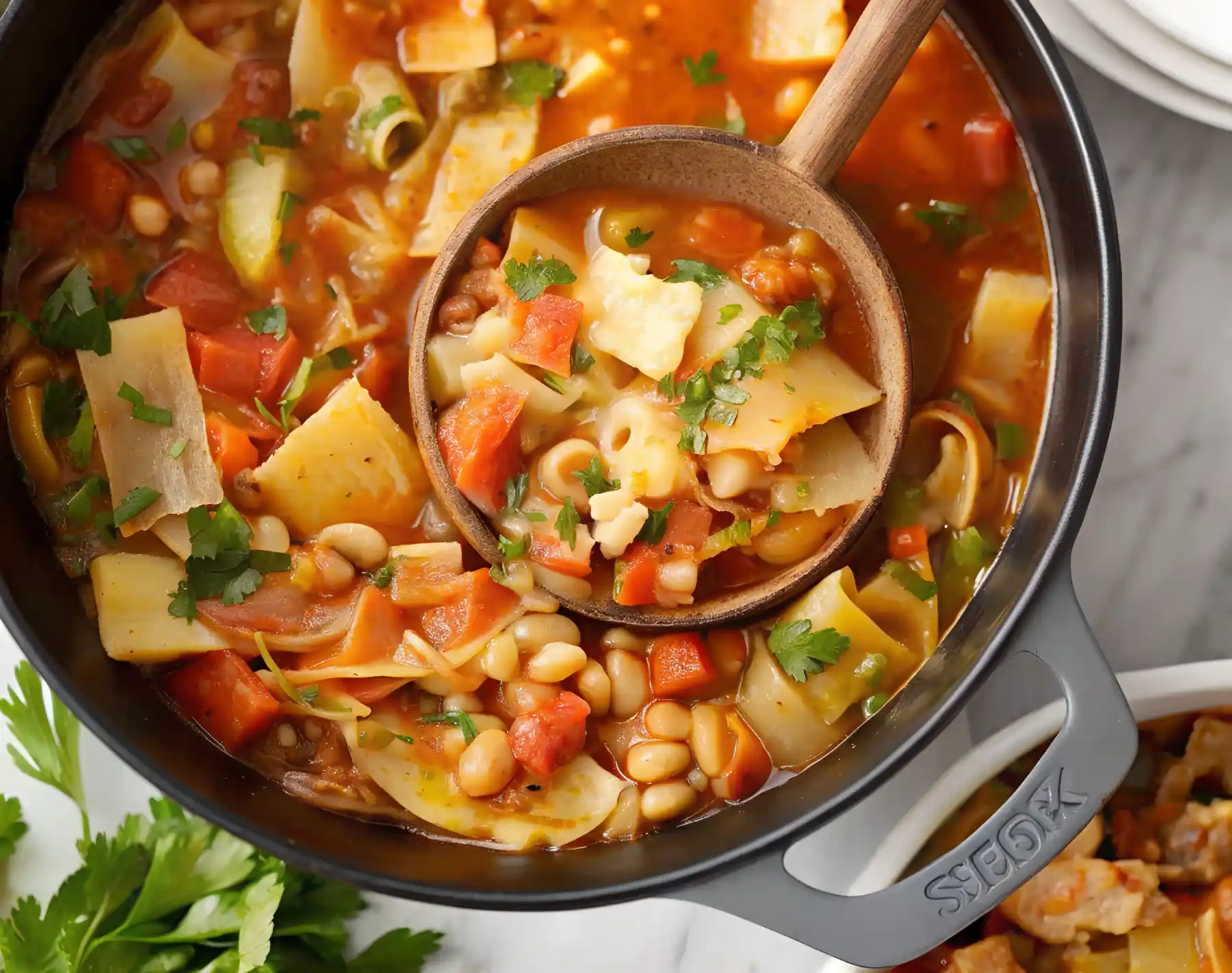 Hearty cabbage soup with pasta and white beans in a black Dutch oven, topped with chopped parsley and served with a wooden ladle.