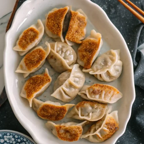 Pan-fried dumplings arranged on a white ceramic plate with crispy golden bottoms and pleated edges.