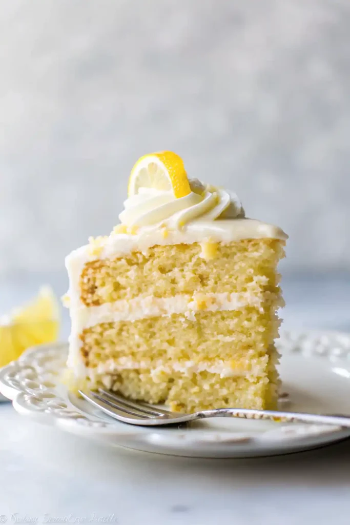 Slice of lemon cake with creamy frosting and a lemon wedge on top.