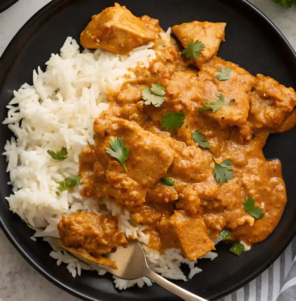 Butter chicken served over steamed white rice on a black plate, garnished with fresh cilantro and a spoon ready to eat.
