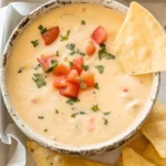 Creamy queso dip in a rustic bowl topped with diced tomatoes and cilantro, served with tortilla chips on the side.