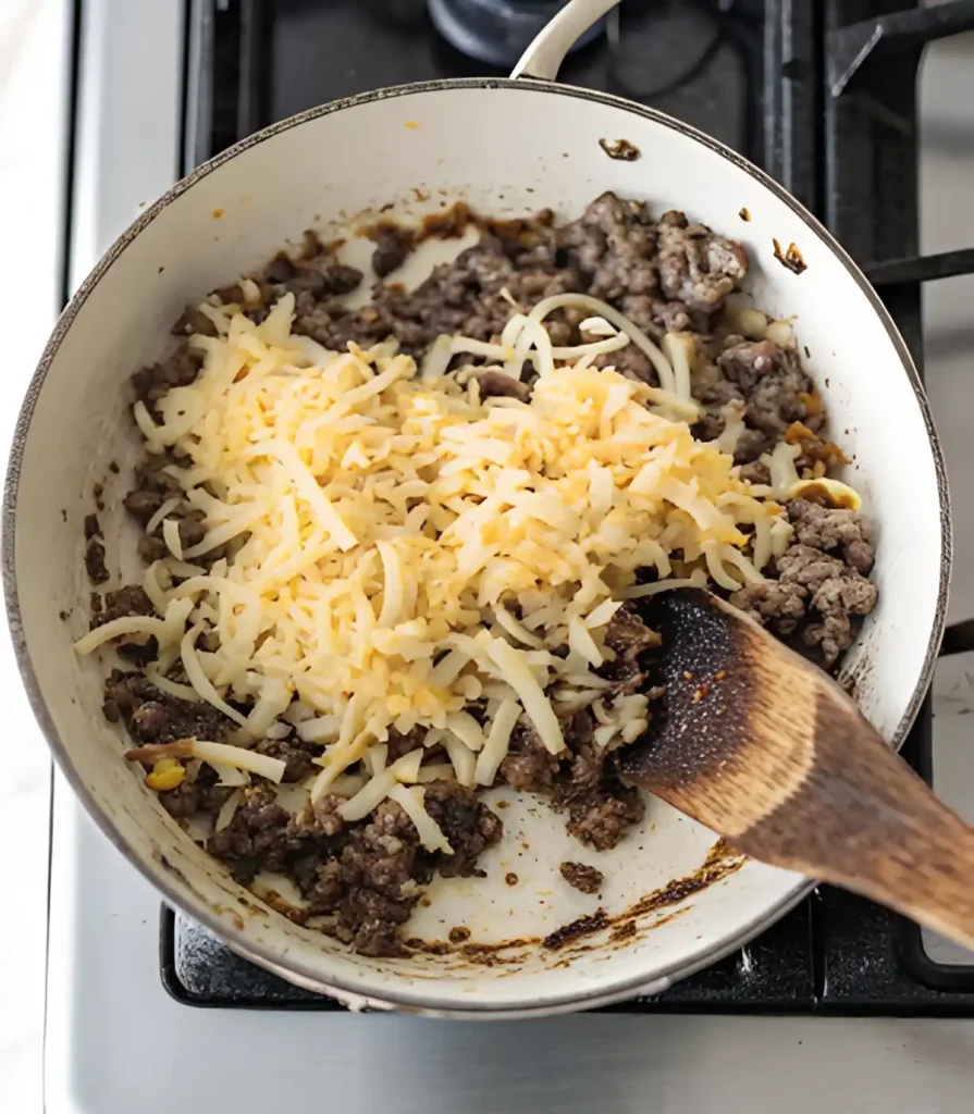 Ground beef cooking in a skillet topped with shredded cheese and stirred with a wooden spoon.