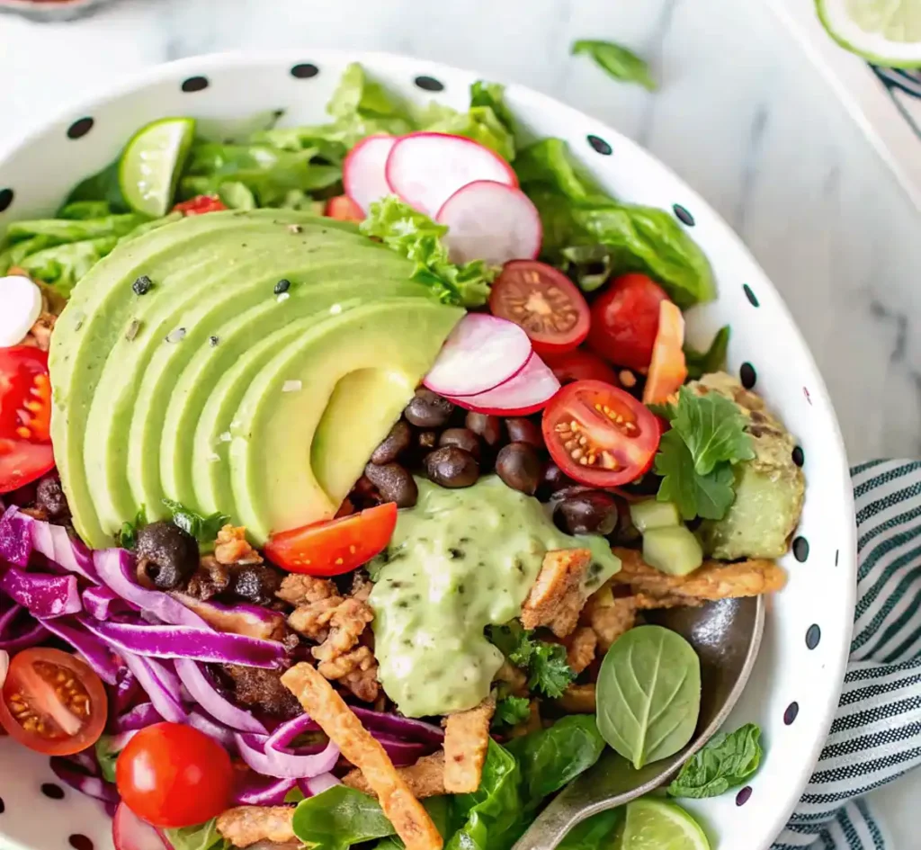 Fresh taco salad in a white bowl with black dots, topped with sliced avocado, cherry tomatoes, radishes, black beans, red cabbage, greens, and creamy cilantro lime dressing.