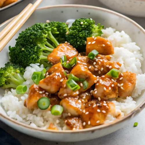 Bowl of teriyaki chicken served over white rice with steamed broccoli, topped with sesame seeds and chopped green onions.