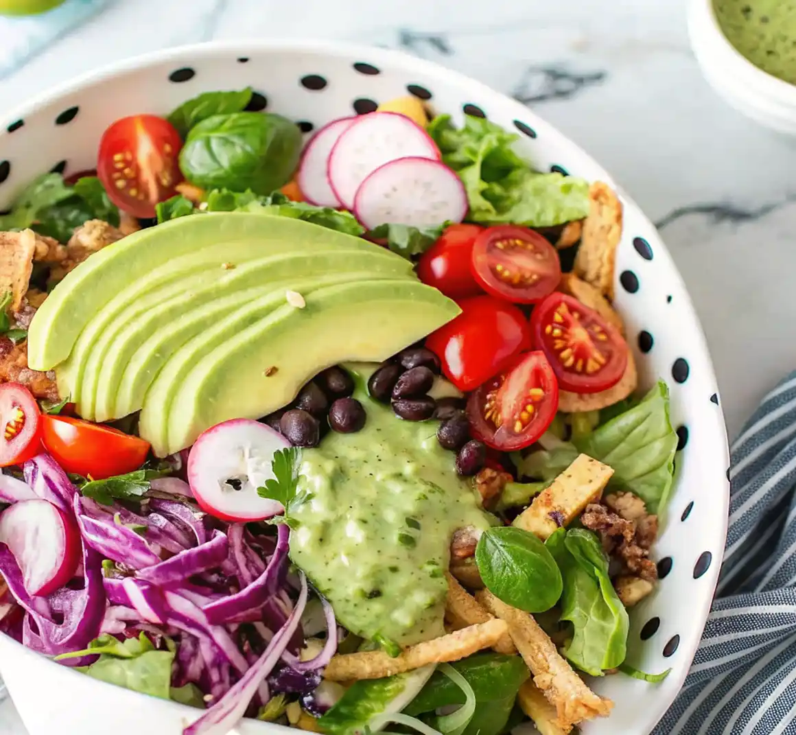 A vibrant taco salad in a white bowl with black dots, topped with avocado slices, cherry tomatoes, radishes, black beans, cabbage, tortilla strips, and creamy cilantro lime dressing.
